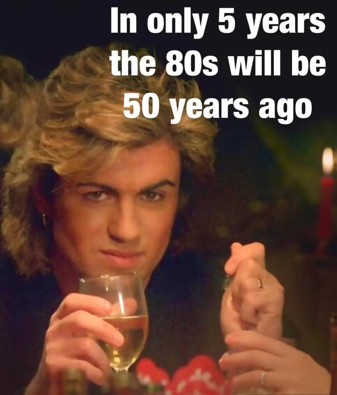 A man with voluminous 80s-style blonde hair holds a glass of champagne while looking intensely at the camera, with a candle…