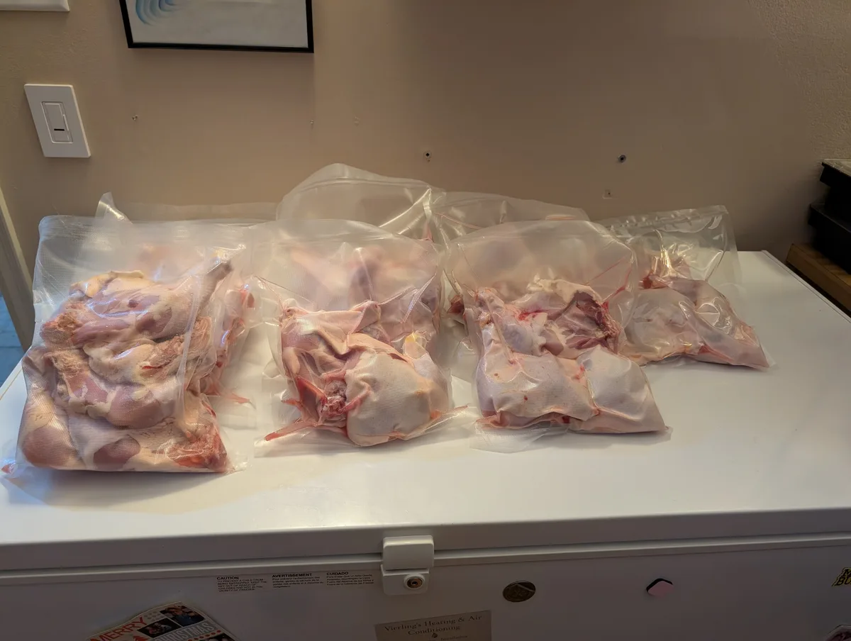 Several vacuum-sealed bags filled with raw chicken parts are arranged on top of a white chest freezer. The freezer has a…