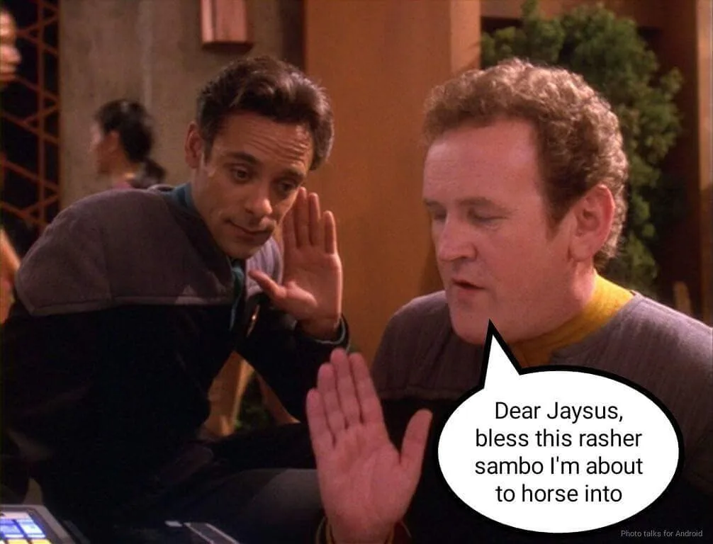 Two men in Star Trek uniforms sit at a table, one with a raised hand in a 'stop' gesture while the other looks down. A…
