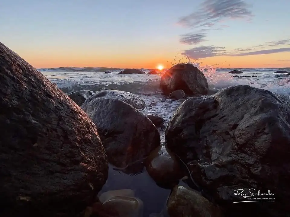 A serene sunset photo of waves crashing against large rocks at the shoreline, with the sun low on the horizon casting orange…