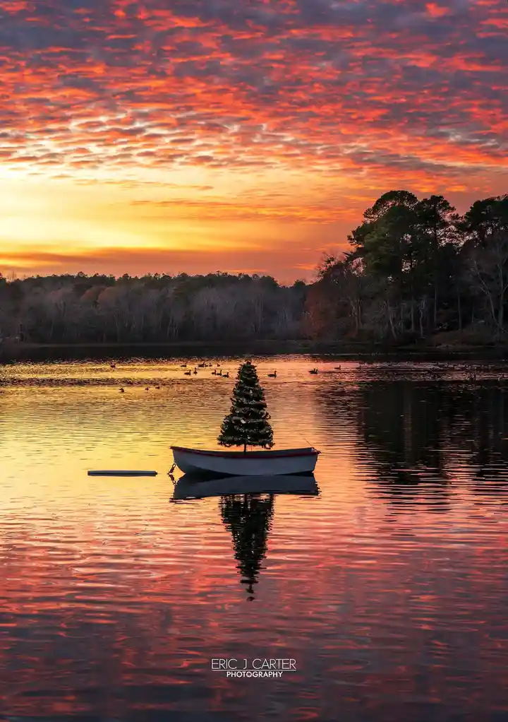 A small white boat floats on a calm lake under a vibrant orange and red sunset sky, with a conical evergreen tree planted in…