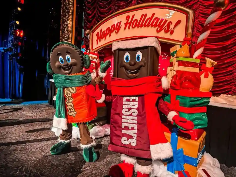 Two Hershey's mascots, a chocolate bar and a cookie, dressed in winter holiday outfits with scarves and hats, stand beside a…