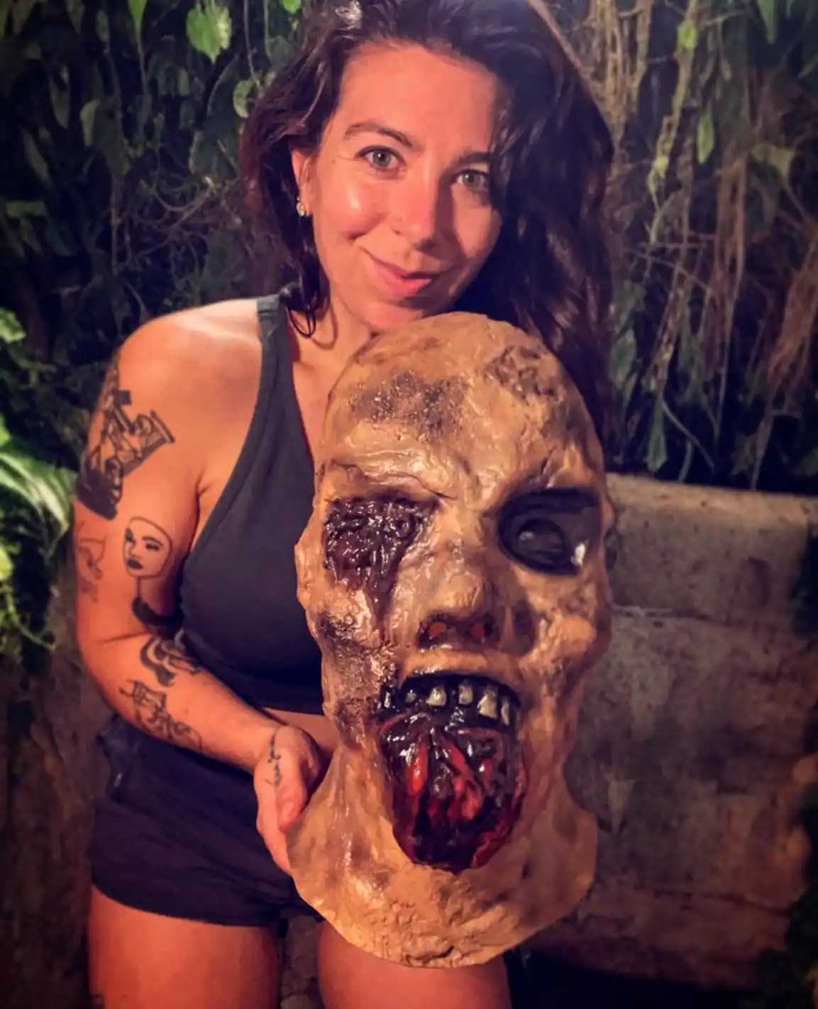 A woman with tattoos on her arms and wearing a black tank top and shorts stands behind a large, gruesome zombie mask with…
