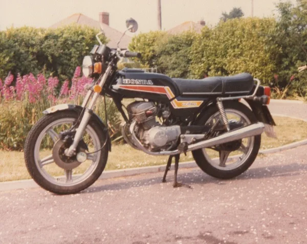 A vintage black Honda CB750 motorcycle is parked on a paved driveway with a hedge and pink flowers in the background. The…