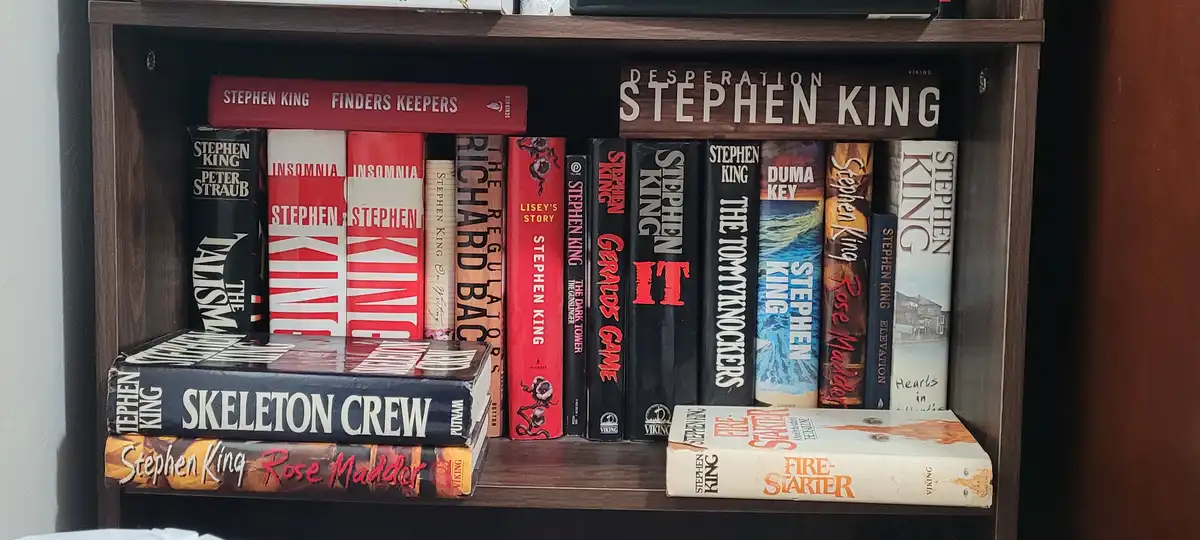 A bookshelf filled with Stephen King novels, including titles like 'It', 'The Dark Tower', and 'Firestarter', with a…