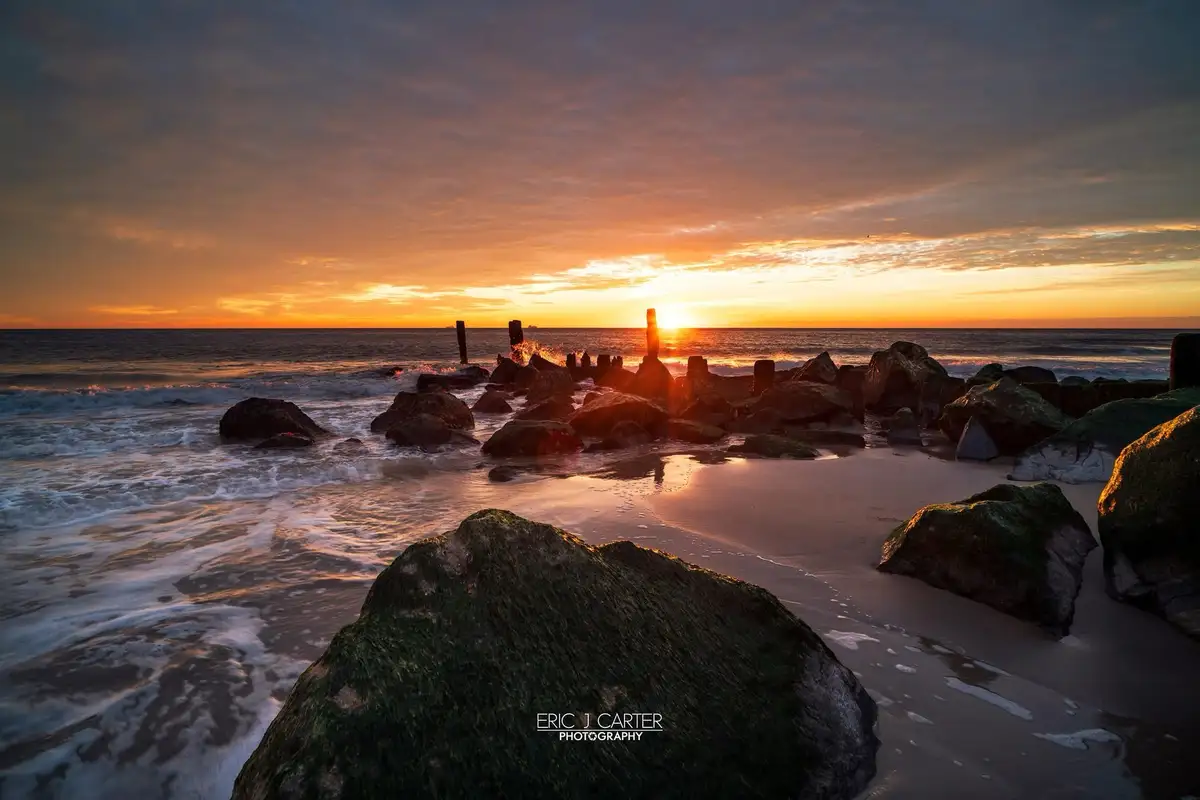 A serene sunset photograph shows waves gently crashing over moss-covered rocks on a beach, with the sun setting on the…