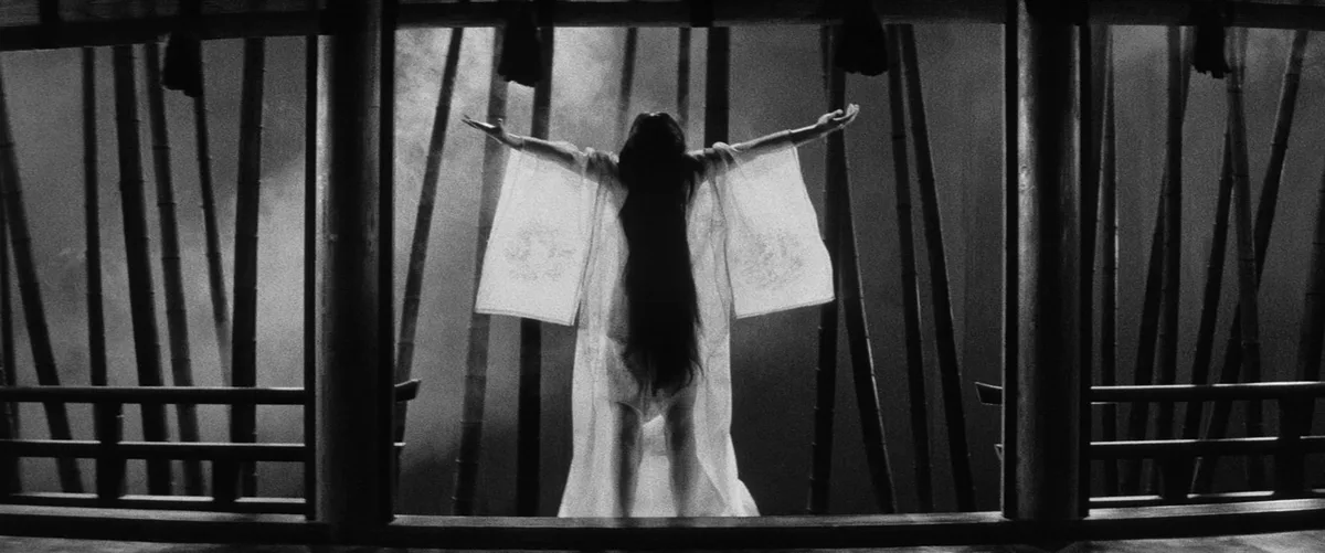 A black-and-white image shows a person with long dark hair, seen from behind, standing on a wooden balcony with arms…