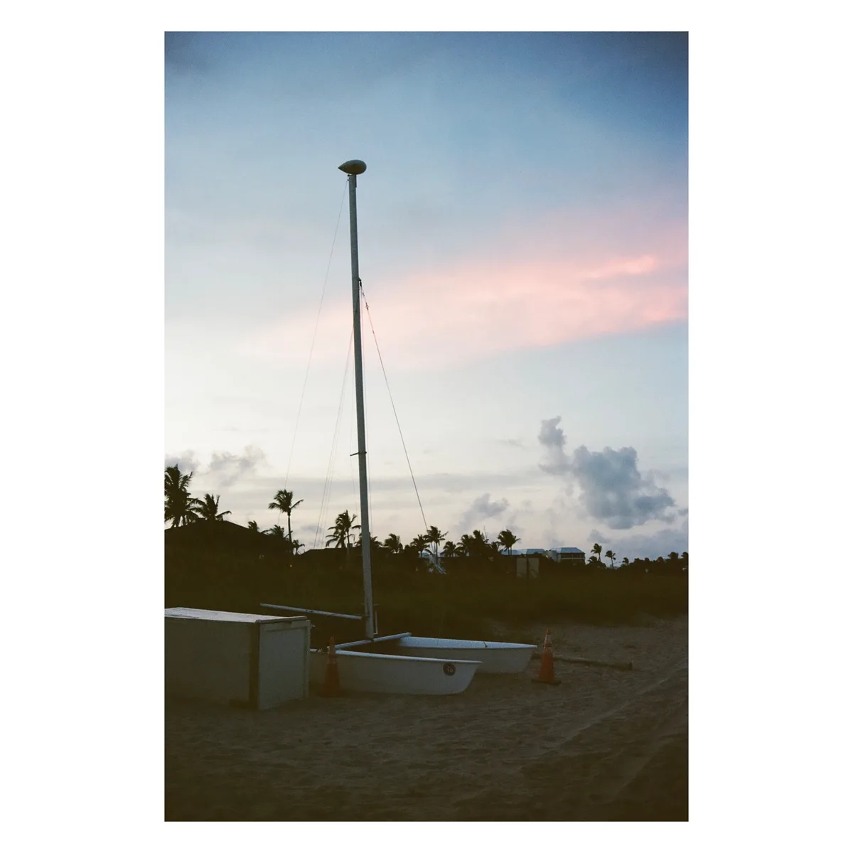 A serene beach scene at dusk with a sailboat's mast standing tall on the sand, flanked by two small white boats and orange…
