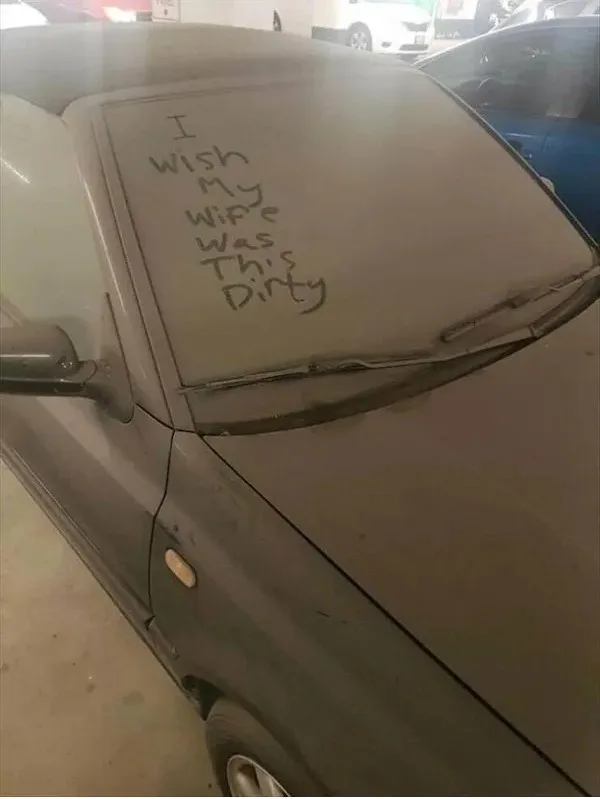 A dirty, dusty gray car is parked in a garage, with handwritten text on its windshield reading 'I wish my wife was this…