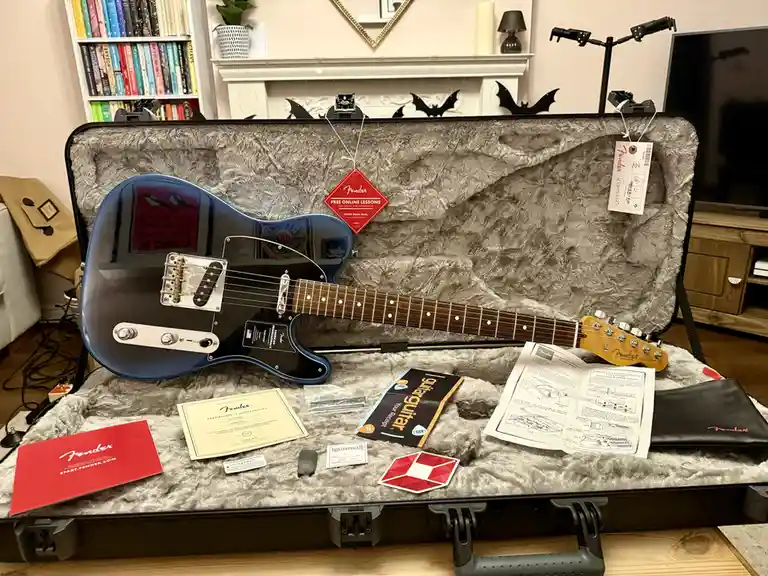 A blue Fender Telecaster electric guitar sits inside its open, plush-lined case, surrounded by official Fender paperwork, a…