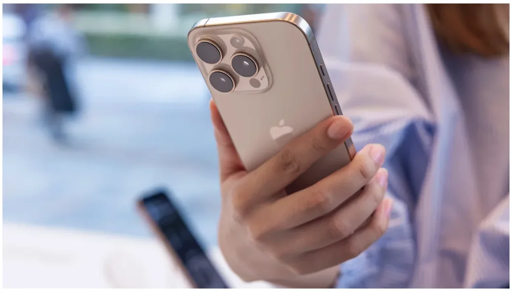 A person holds a gold-colored iPhone with a prominent triple-camera system, looking at it intently. The overlaid text reads…