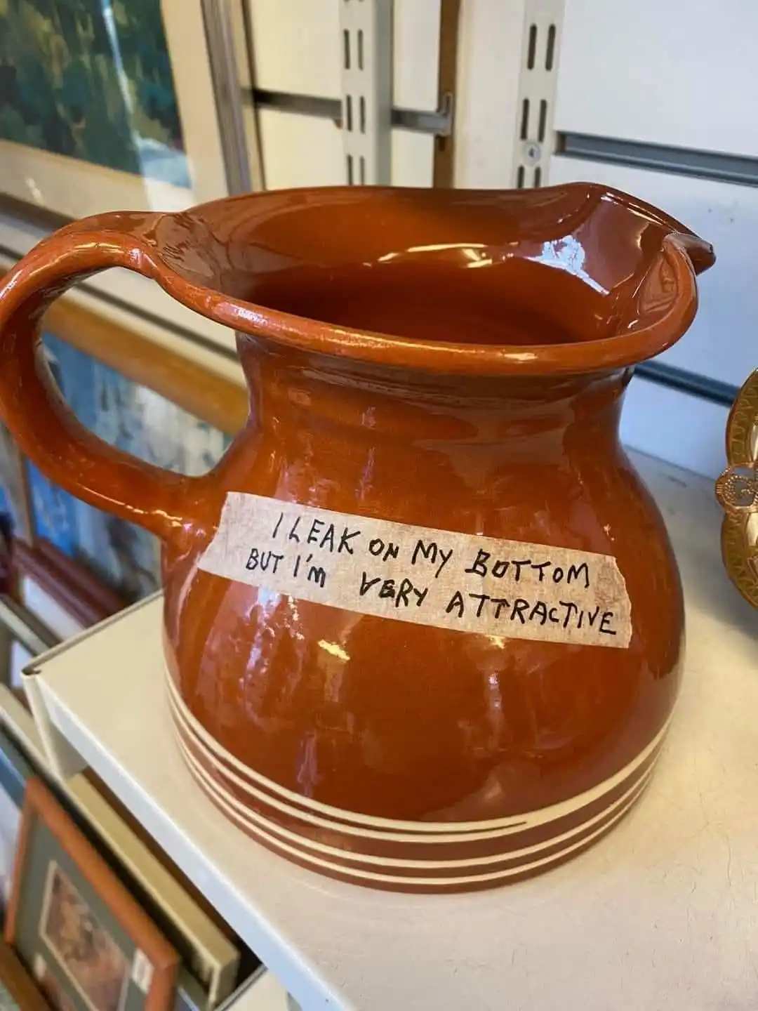 A glossy, terracotta-colored ceramic pitcher with a handle sits on a white shelf, featuring a handwritten note taped to its…
