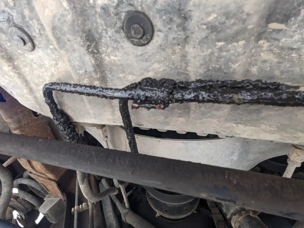 A close-up photo of a severely corroded and oil-slicked metal pipe under a vehicle, with visible rust and grime. The…