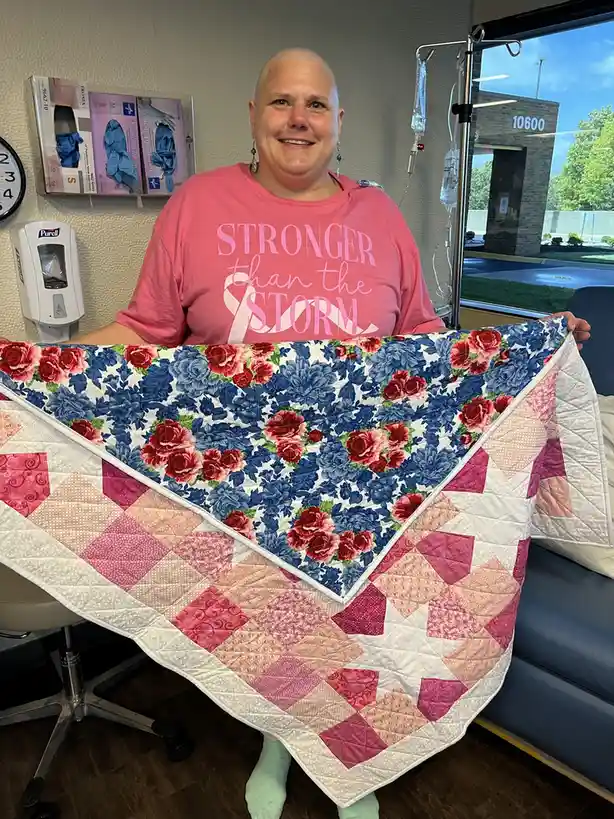 A bald woman in a pink t-shirt with the text 'STRONGER than the STORM' and a pink ribbon graphic smiles while holding a…