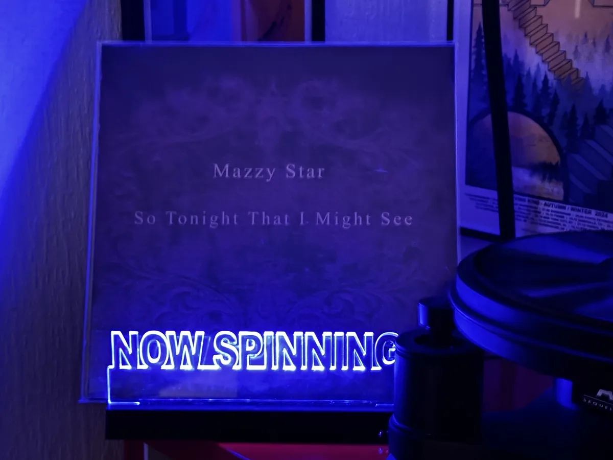A vinyl record sleeve for Mazzy Star's album 'So Tonight That I Might See' is shown under blue light, with a glowing neon…