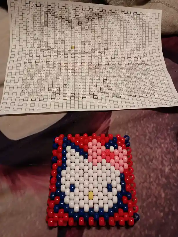 A handmade beaded craft, likely a bracelet or keychain, featuring a pattern of red, white, blue, and a single yellow bead…