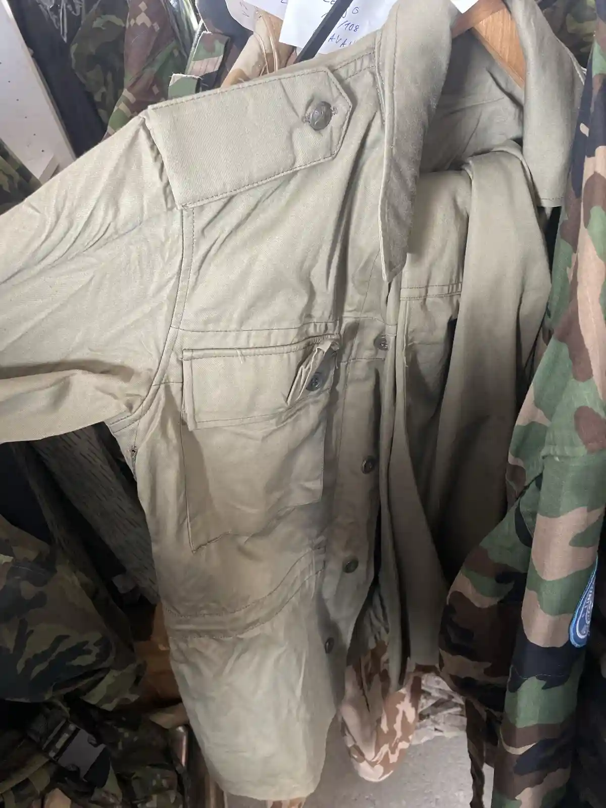 A beige military-style jacket hangs on a wooden hanger among other camouflage-patterned uniforms in what appears to be a…