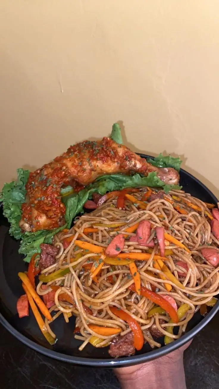 a hand holds a black plate with a large serving of stir-fried noodles mixed with sliced carrots, bell peppers, and chunks of…
