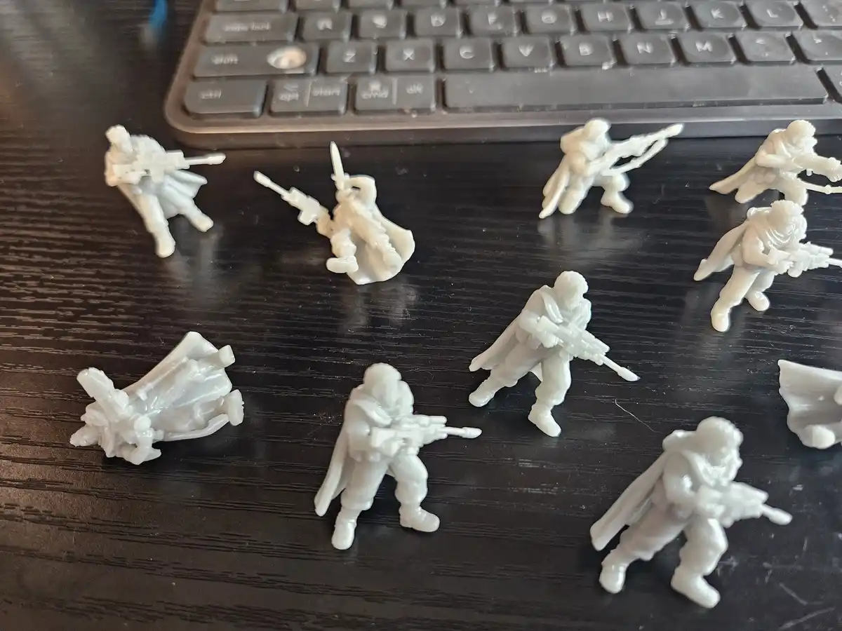 Several white 3D-printed miniature soldier figures with guns and capes are scattered on a dark wooden desk in front of a…