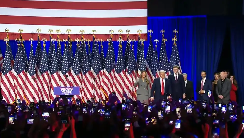 A wide shot of Donald Trump, Melania Trump, and other family members standing on a stage in front of a large American flag…