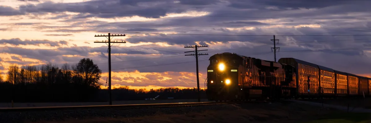 A train with the number 5449 illuminated on its front is moving toward the viewer at dusk, its headlights glowing against a…