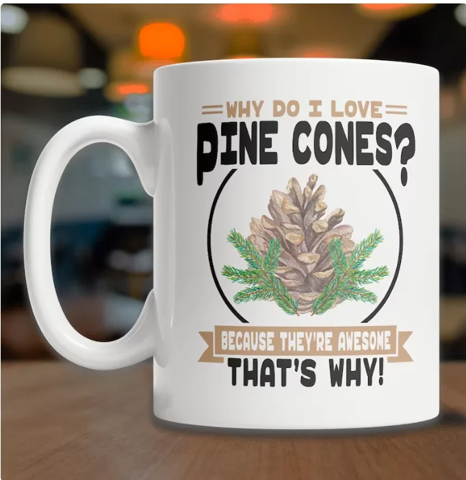 A white ceramic mug sits on a wooden surface, featuring a printed design with a pine cone and pine needles inside a circle…