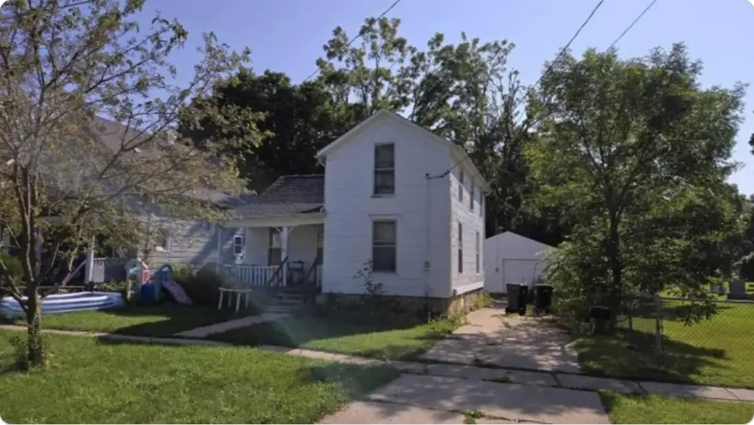 a photo of a modest white two-story house with a small front yard, surrounded by trees and a concrete walkway, with…