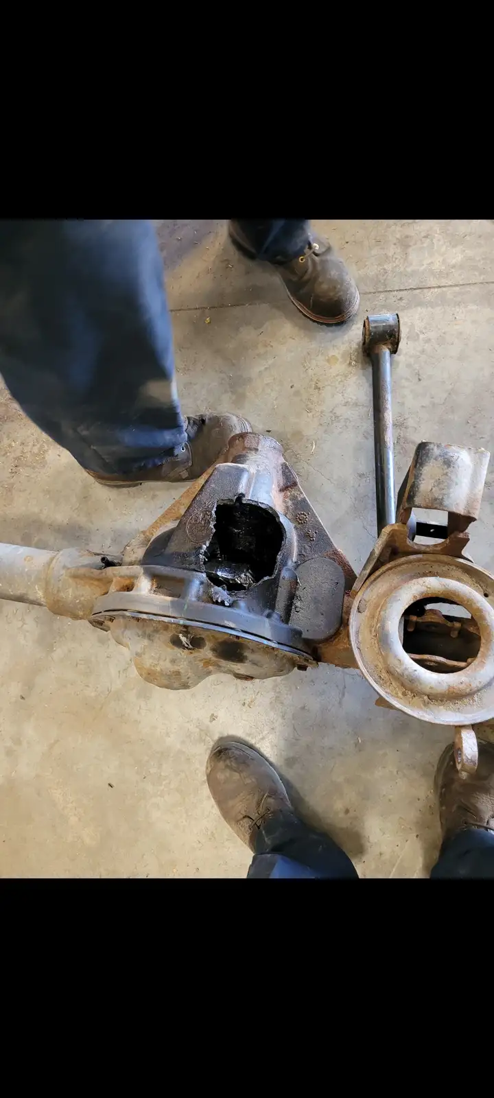 A top-down view shows a person's dirty work boots standing around a severely damaged, open-ended automotive differential…