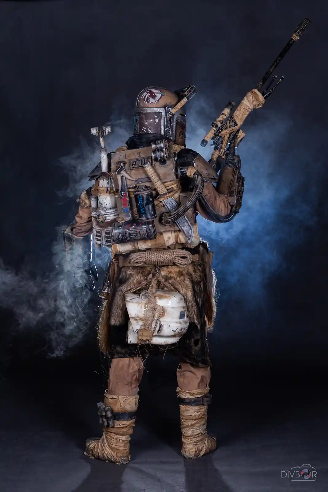 A person in a heavily modified, weathered Mandalorian armor costume stands in a smoky studio, holding a blaster rifle. The…