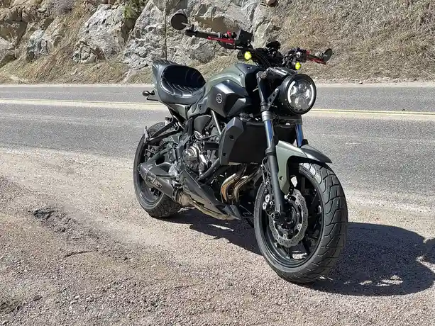 A matte olive-green Yamaha FZ-07 motorcycle is parked on a gravel shoulder beside a paved road, with a rocky, dry hillside…