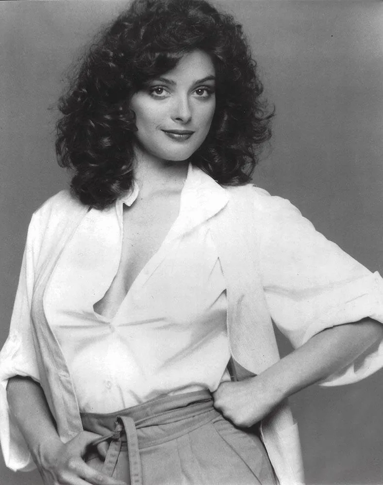 A black-and-white photo of a woman with voluminous curly hair, wearing a white button-up shirt that is unbuttoned low and…