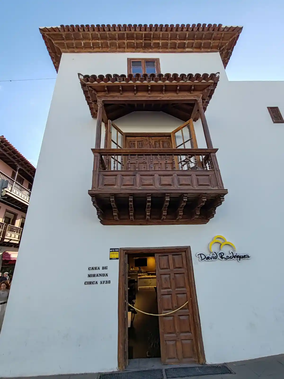 A white building with a traditional wooden balcony and door, labeled 'CASA DE MIRANDA CIRCA 3730' on the wall, humorously…