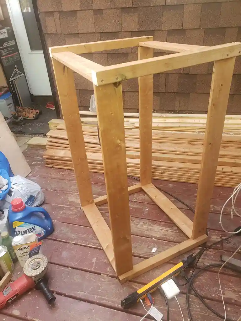 A partially assembled wooden frame, resembling a table base, sits on a red wooden deck surrounded by construction tools and…