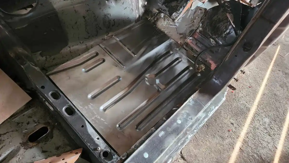 A close-up photo shows the bare metal floor of a vehicle's interior, with visible rust, grime, and exposed structural…