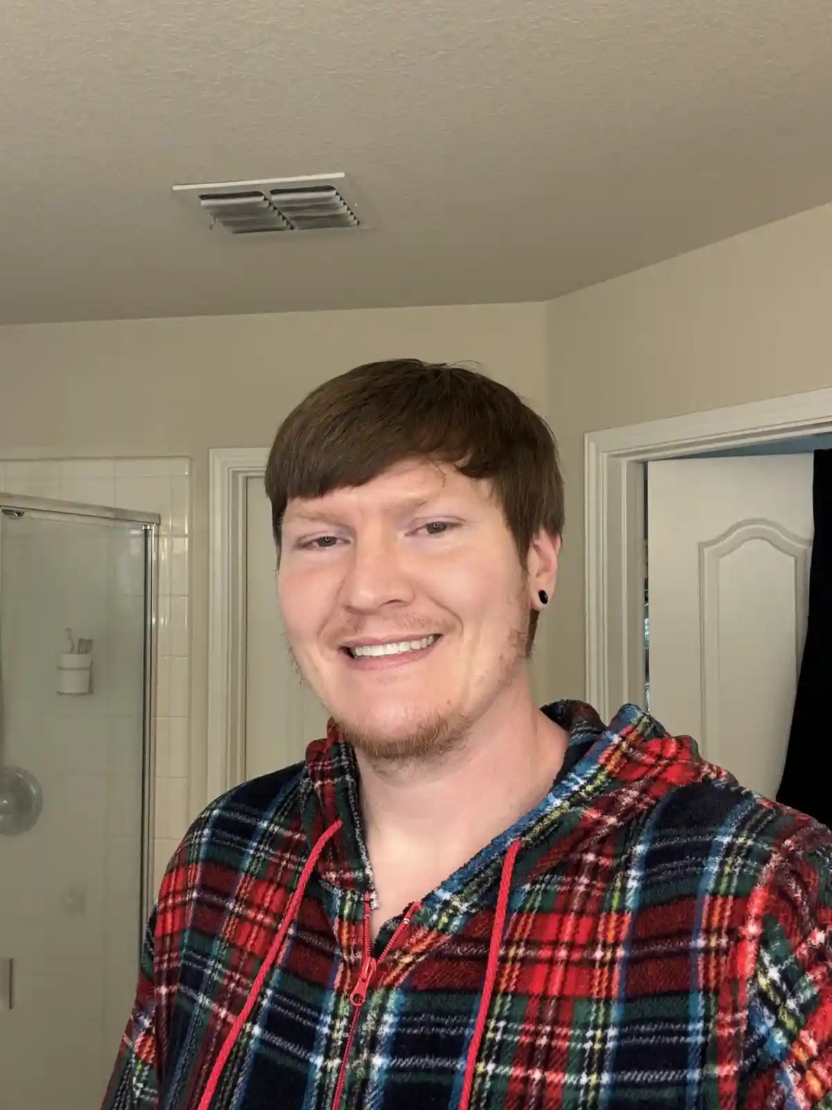 A man with brown hair and a goatee, wearing a red, green, and black plaid zip-up hoodie with red drawstrings, smiles at the…