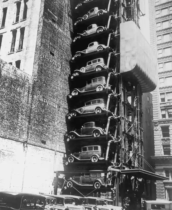 a black-and-white historical photograph shows a tall, narrow vertical parking structure attached to a brick building…