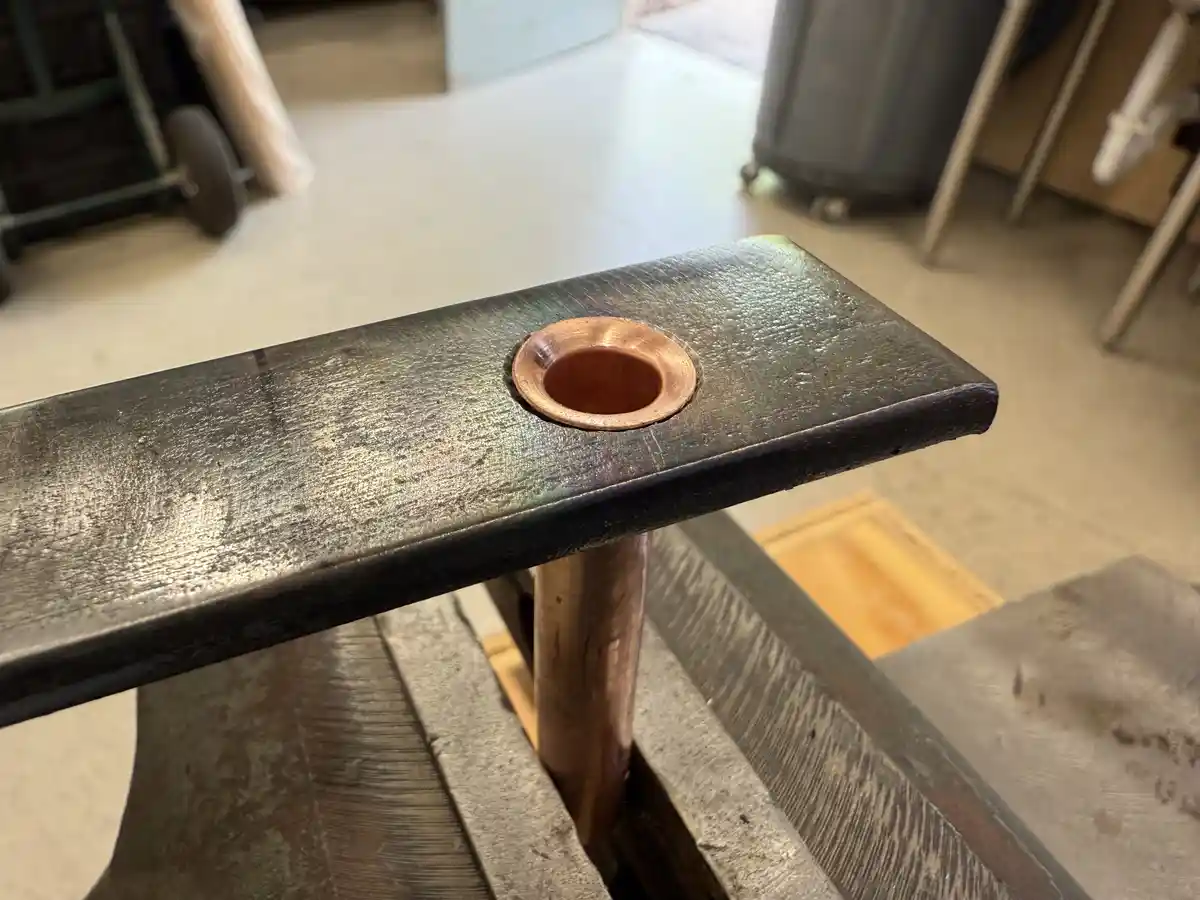 A close-up photo shows a shiny copper pipe inserted into a dark, worn metal workbench or table. The pipe protrudes…