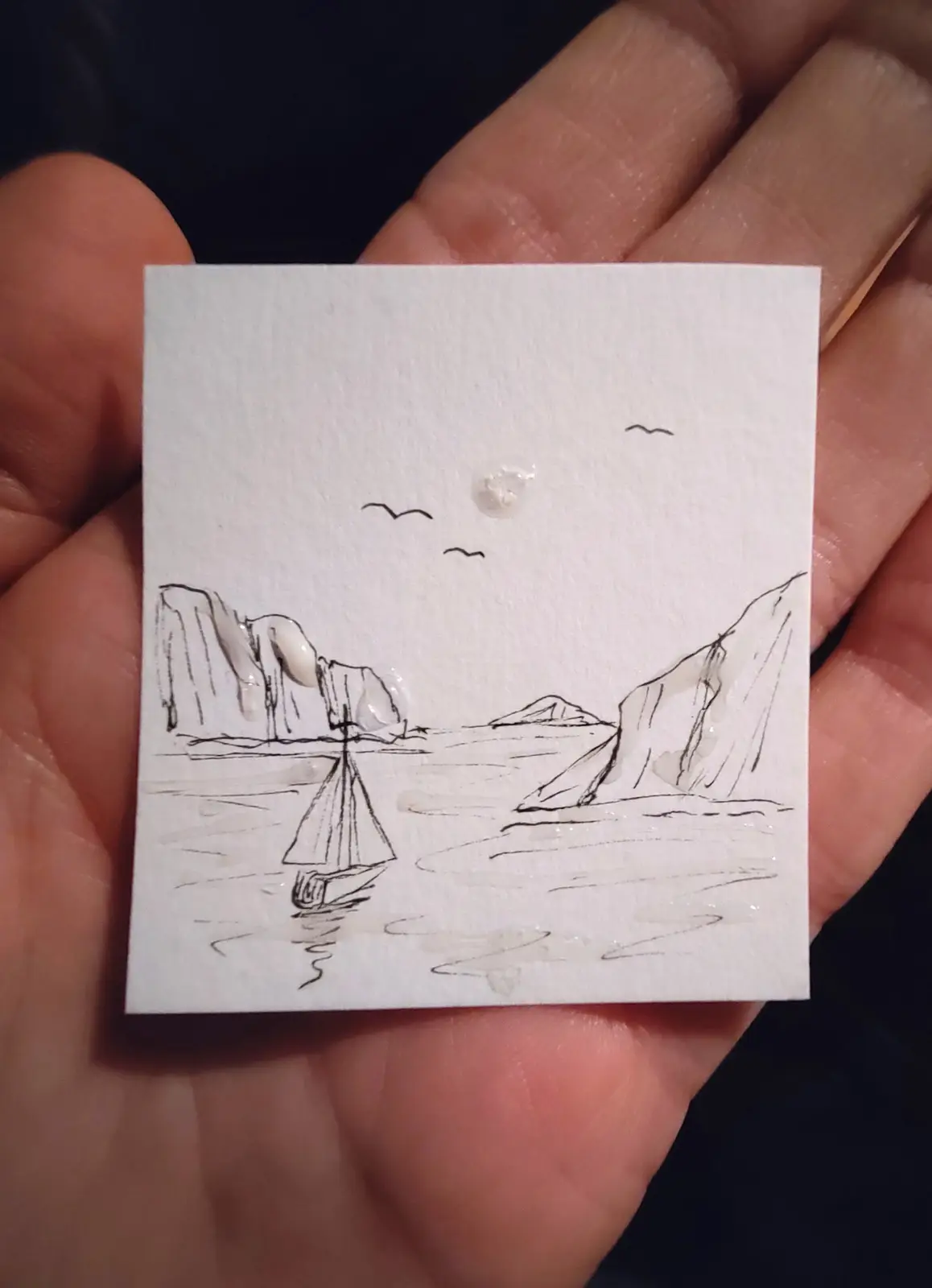 A hand holds a small square of white paper with a simple black ink sketch of a sailboat on water between two cliffs, with…