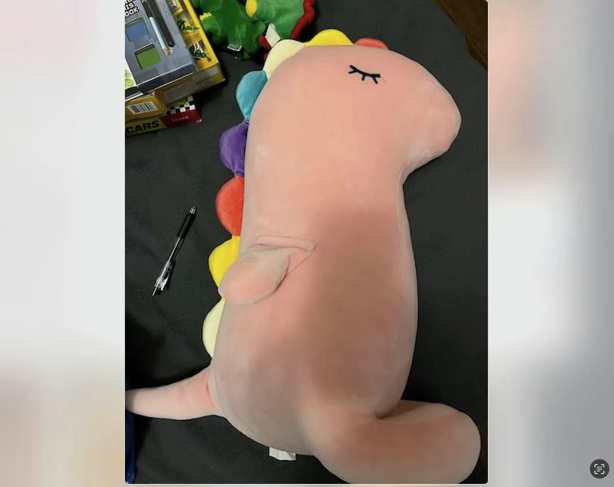 A large, pink plush dinosaur with a rainbow-colored spine is lying on its back on a dark surface, appearing to be sleeping…