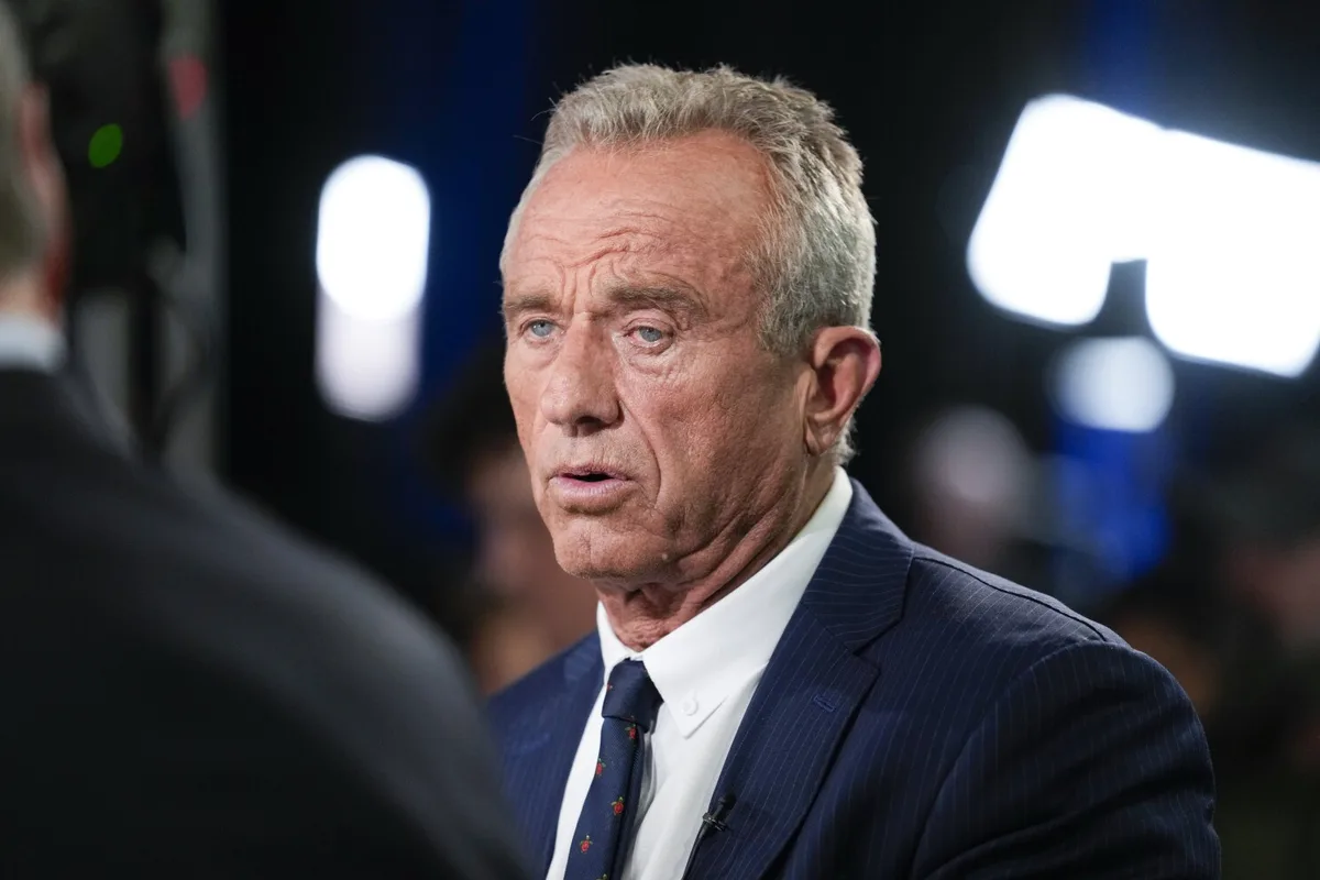a close-up photo of an older man with gray hair, wearing a dark pinstripe suit and tie, looking surprised or confused while…
