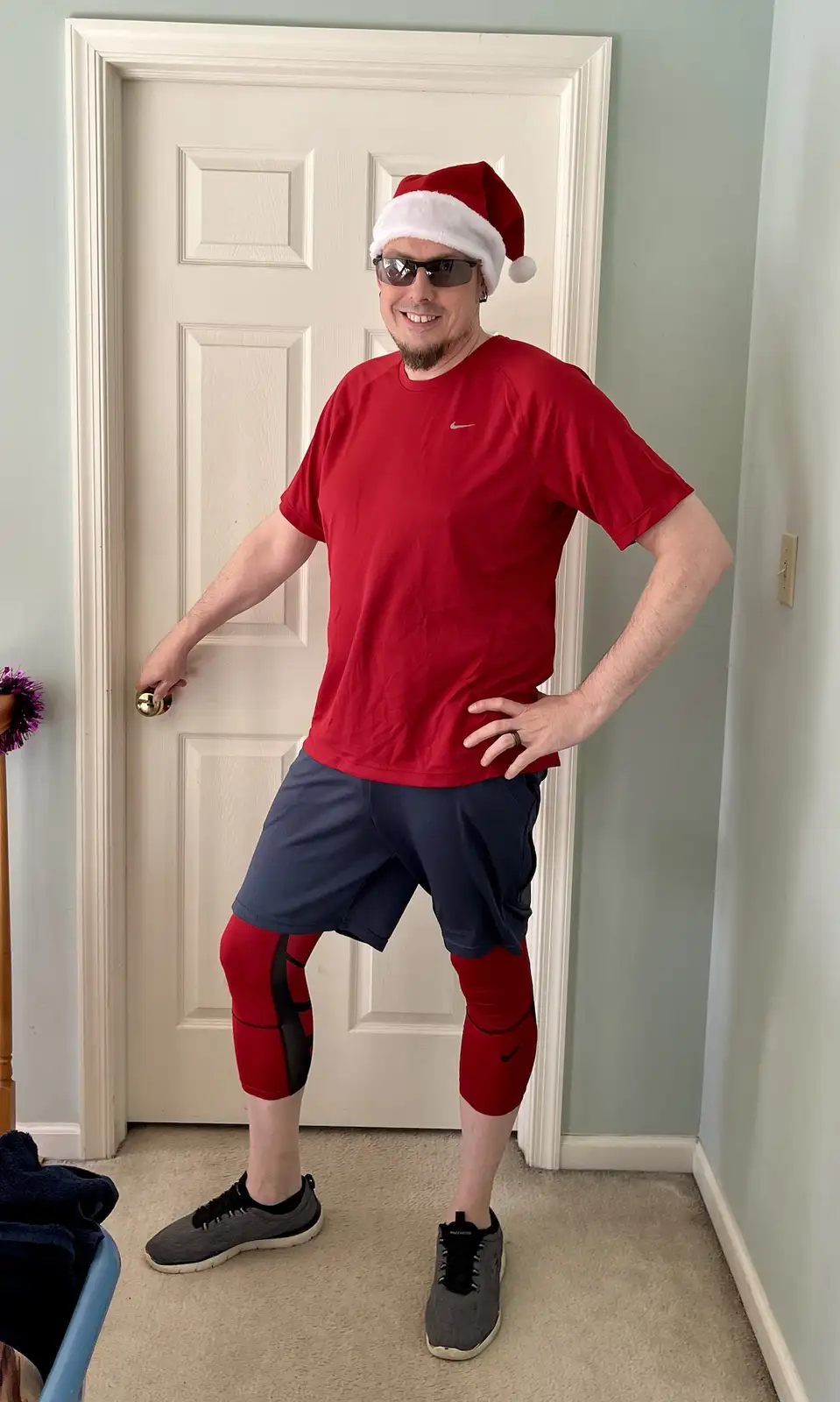 A man in athletic wear, including a red t-shirt, blue shorts, and red compression leggings, stands in a hallway in front of…