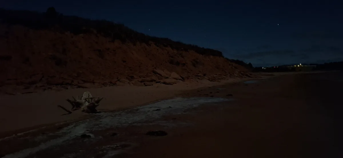 A dark, nighttime beach scene with a sandy shore, a rocky dune in the background, and faint lights from distant buildings. A…