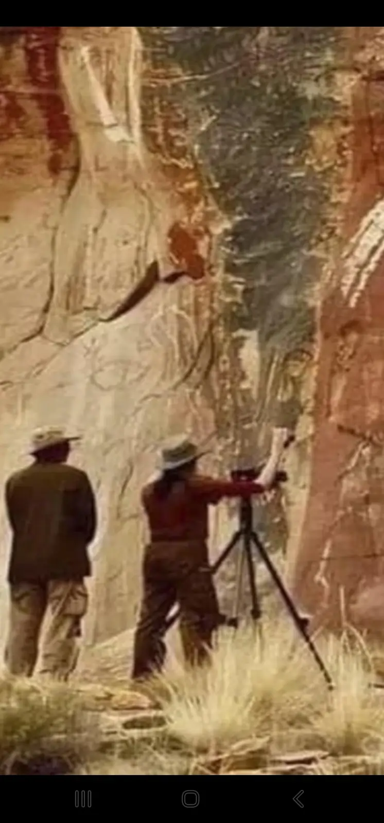 Two people stand before a large, textured rock face with ancient-looking carvings. One person, wearing a hat, is operating a…