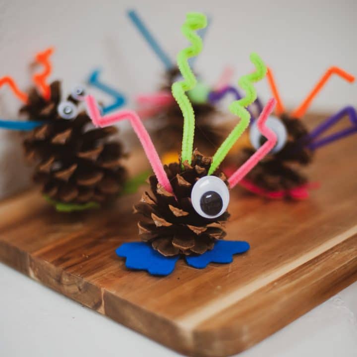 Several pinecones are decorated as whimsical monsters with googly eyes, colorful pipe cleaner limbs, and blue felt feet…