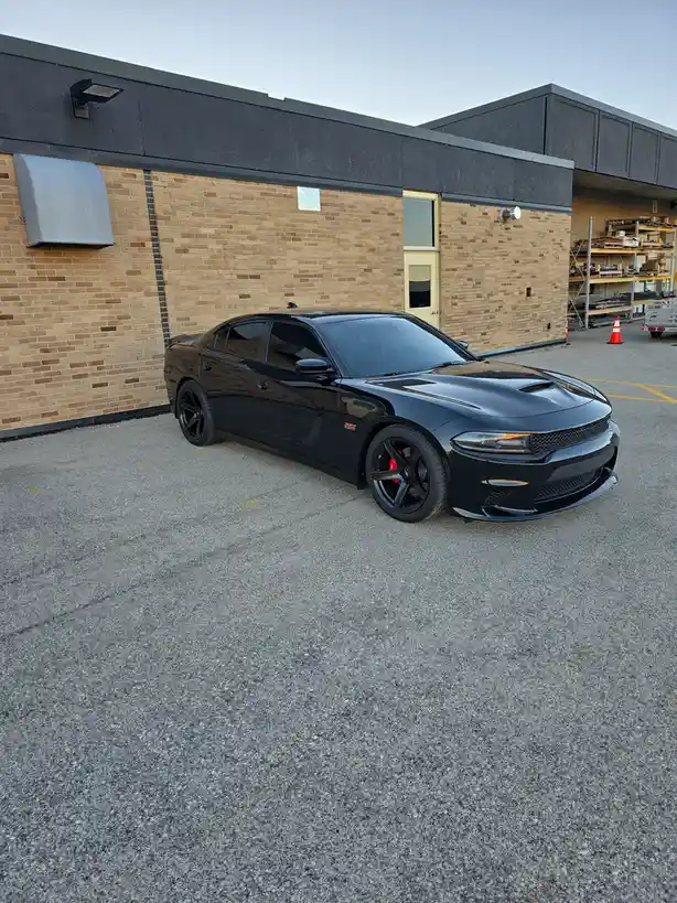 A glossy black Dodge Charger parked in a gravel lot beside a brick and dark panel building. The car features black rims with…