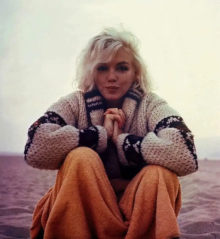 a vintage photograph of Marilyn Monroe sitting on a beach in a cream and black patterned knit sweater and orange pants…