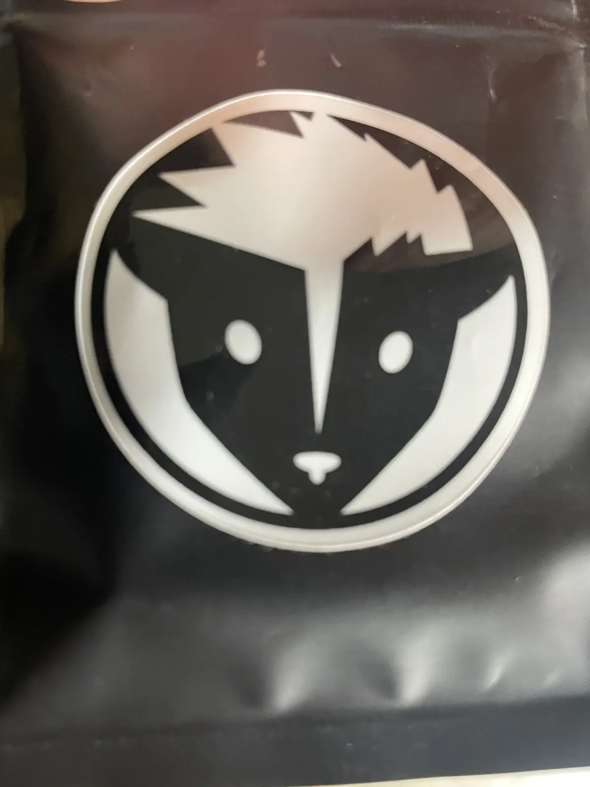 A close-up photo shows a black and white circular sticker of a stylized skunk face with a spiky white topknot, white eyes…