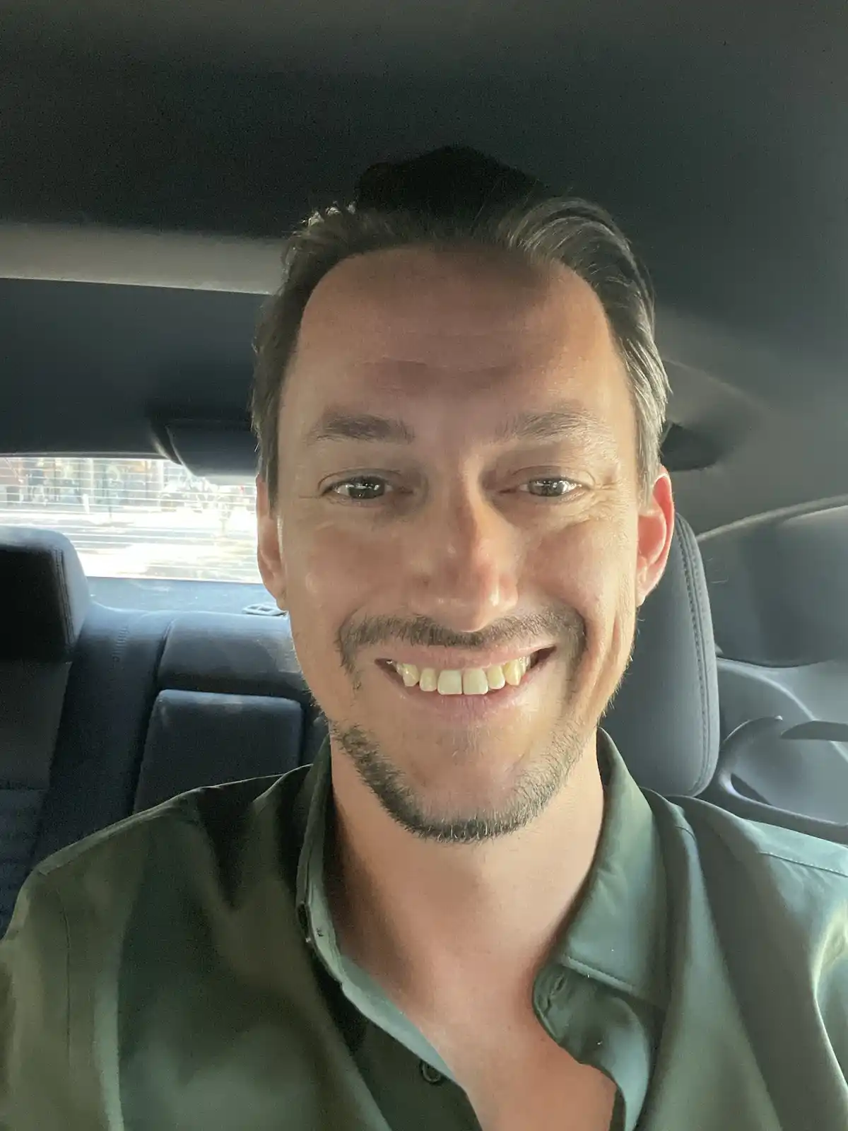 A man with a wide, toothy smile is taking a selfie from inside a car, showing his teeth which appear slightly yellowed. He…