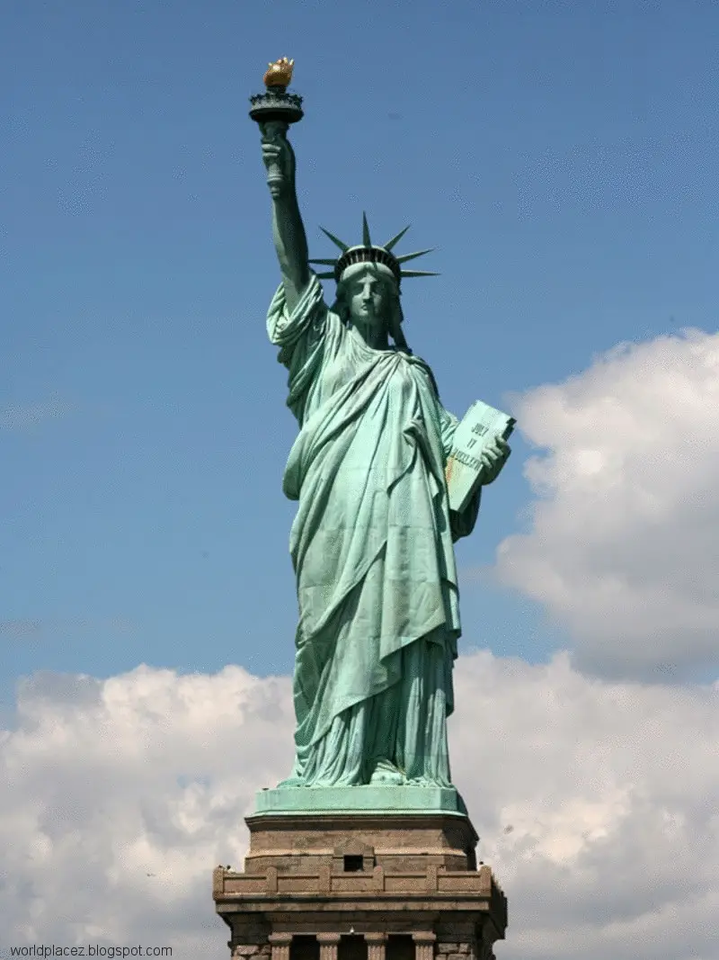 The image shows the Statue of Liberty against a blue sky with white clouds, holding her torch aloft and a tablet in her…