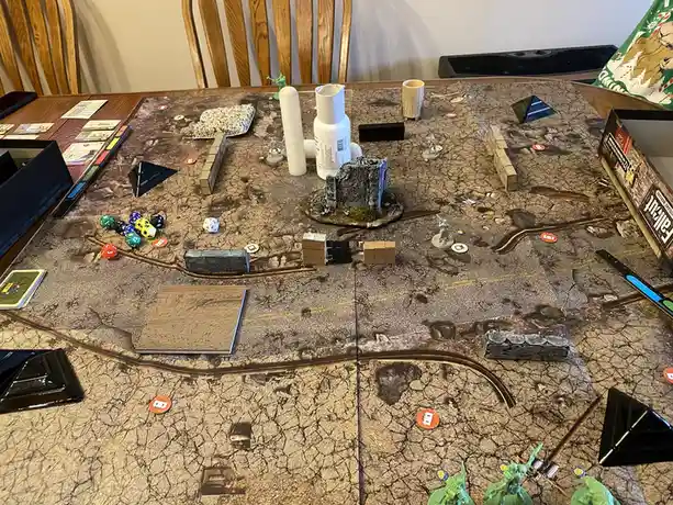An overhead shot of a tabletop wargame setup featuring a cracked desert map, miniature figures, dice, cardboard terrain…